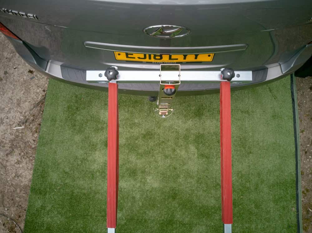 Alt text: Car mounted on a bike rack with red support arms, ready for transport, on a green artificial grass surface, showcasing triathlon bike transportation setup.