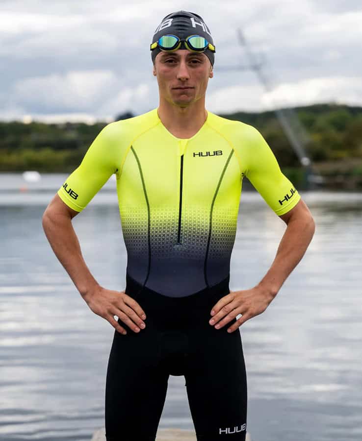 Triathlon athlete wearing yellow and black HUUB wetsuit and swim cap near water during training or race.