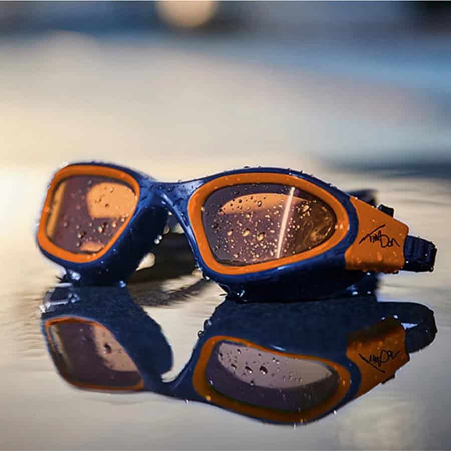 A pair of blue and orange triathlon goggles with water droplets on the lenses, reflecting on a wet surface, highlighting triathlon gear essentials for open water swimming.