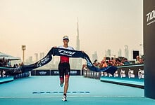 Victory athlete crossing the finish line with the triathlon tape at an international triathlon event in Dubai. Celebrating achievement with Dubai city skyline in the background, promoting triathlon race success.