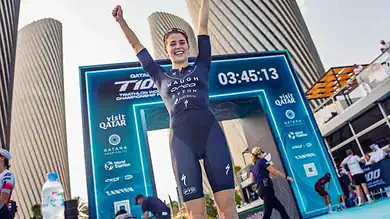 1. Female triathlete celebrating victory at finish line with digital timer, modern city skyline background, and triathlon event branding in Qatar, 2023.