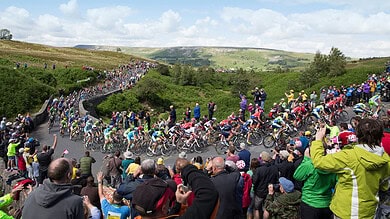 A large group of cyclists competing in a triathlon race through a scenic, lush countryside with enthusiastic spectators cheering on the event.