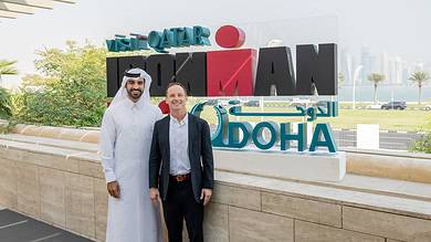 1. Diverse men smiling at Doha international triathlon event, wearing traditional and business attire, with iconic Doha skyline and Qatar sign in background.