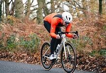 An athlete in a red and black cycling kit with a white helmet riding a road bike through a forested area during autumn.