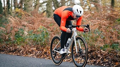 An athlete in a red and black cycling kit with a white helmet riding a road bike through a forested area during autumn.
