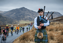 Piper dressed in traditional Scottish attire playing bagpipes while cyclists pass by on a rural road with mountains in the background.