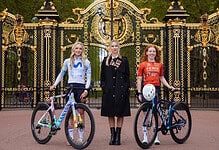 Three female cyclists with bikes standing in front of ornate royal park gates, dressed in athletic gear, ready for a triathlon training session or cycling event.