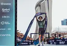 An athlete crosses the finish line at a triathlon event in Singapore, holding the finisher tape high in victory. The iconic Marina Bay Sands skyline is visible in the background, highlighting the even.