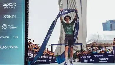 An athlete crosses the finish line at a triathlon event in Singapore, holding the finisher tape high in victory. The iconic Marina Bay Sands skyline is visible in the background, highlighting the even.