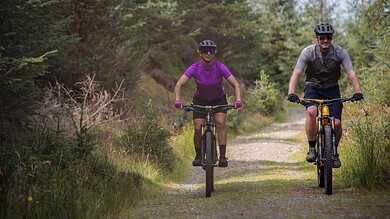 Two cyclists riding mountain bikes on a forest trail during daytime, surrounded by lush green trees and nature.