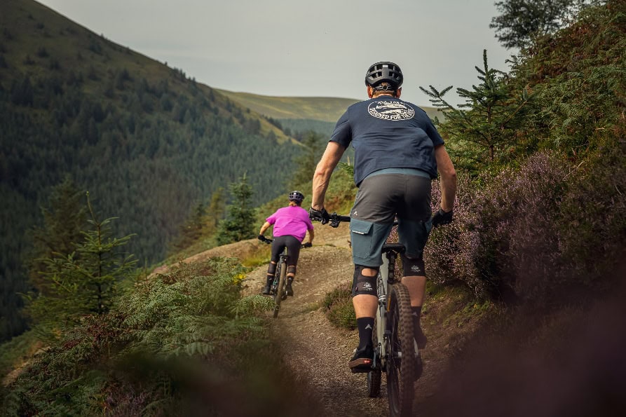 Two cyclists riding mountain bikes on a trail through lush green hills and forested landscape, showcasing outdoor adventure and fitness.
