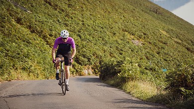 An experienced cyclist wearing a purple jersey and helmet riding along a winding mountain road surrounded by lush greenery and hills, showcasing outdoor endurance training.