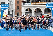 Triathlon athletes start race in urban city setting, walking with bikes on blue carpet, with spectators and historic buildings in background.