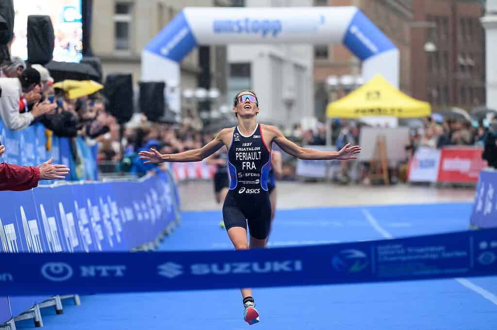Female triathlete crossing the finish line with arms outstretched, celebrating her achievement at a triathlon event, with spectators and race banners in the background.