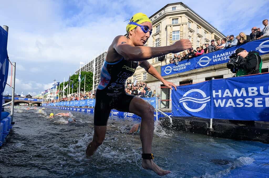Triathlon athlete emerging from water during race, wearing wetsuit, yellow swim cap, and goggles, with spectators and race banners in the background.