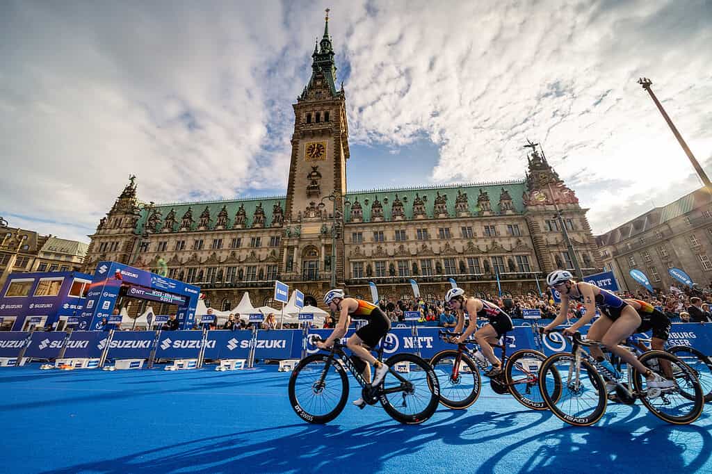 Professional triathletes cycling past the historic city hall during a triathlon event in a European city, with spectators and race banners in the background.
