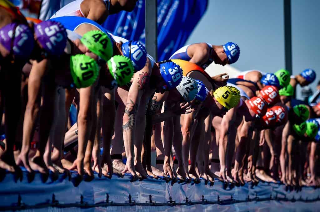 Athletes in colorful gear and helmets at the starting line of a triathlon race, ready to begin the swim segment.
