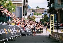 A detailed view of a triathlon finish line with athletes crossing, surrounded by cheering spectators and modern urban architecture, highlighting endurance and competitive spirit.