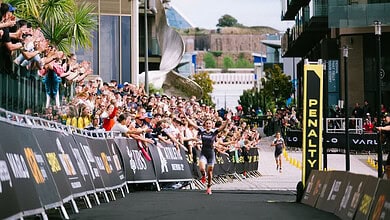 A detailed view of a triathlon finish line with athletes crossing, surrounded by cheering spectators and modern urban architecture, highlighting endurance and competitive spirit.