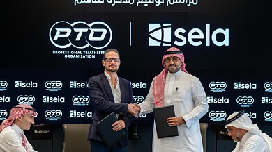 Two men shaking hands during the MOU signing ceremony for triathlon collaboration, with a backdrop displaying logos of PTO and Sela.