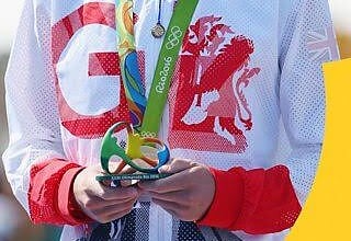 Jonny Brownlee smiling with Olympic gold medal around his neck, wearing Team GB sportswear, during the Olympic medal ceremony.