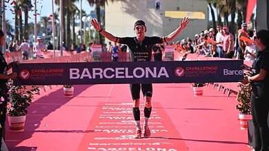 Triathlete crosses the finish line at Challenge Barcelona with arms raised in victory as spectators cheer along a pink finish chute lined with palm trees.