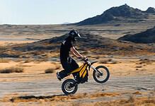 A cyclist wearing protective gear performs a wheelie on a dirt trail in a desert landscape, with rugged hills in the background.