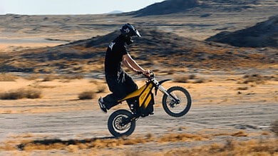 A cyclist wearing protective gear performs a wheelie on a dirt trail in a desert landscape, with rugged hills in the background.