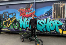 Young man with an electric bike in front of a colourful graffiti mural featuring the words 'Pedal Power Workshop' and a bicycle illustration.