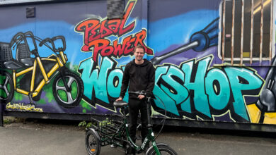 Young man with an electric bike in front of a colourful graffiti mural featuring the words 'Pedal Power Workshop' and a bicycle illustration.
