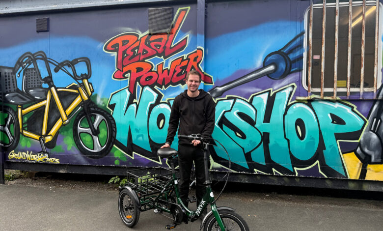 Young man with an electric bike in front of a colourful graffiti mural featuring the words 'Pedal Power Workshop' and a bicycle illustration.