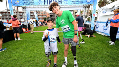 Two young athletes at the finish line of the San Diego Triathlon Challenge, smiling with medals and bib numbers 3009 and 3023, on a grassy field with event banners in the background.