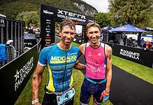 Two male triathletes smiling at the finish line of the Xterra World Championship in Italy, with mountains in the background and race banners around.