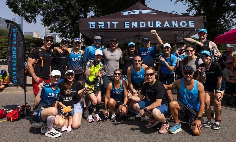 Group of athletes posing for a photo under a GT endurance tent at a race event, wearing blue and black triathlon apparel, sunglasses and caps, sunny day.