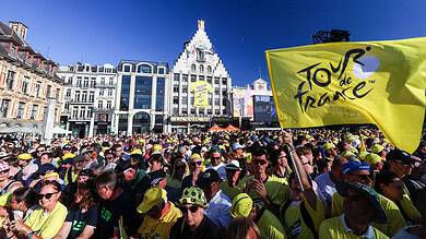 Large crowd of cyclists and spectators at the Tour de France start line in a city square, with a prominent yellow Tour de France flag and historic buildings in the background.