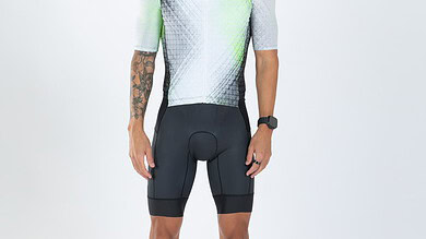 Male triathlete in cycling gear with sunglasses, standing against a white background, showcasing professional triathlon apparel and equipment.