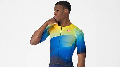 Male triathlon athlete wearing a colourful cycling suit with yellow, blue, and green shades, standing in a studio with a white background.