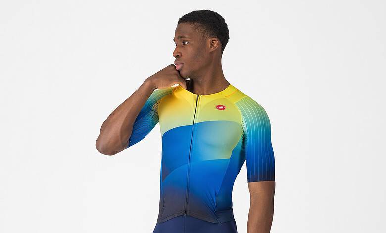Male triathlon athlete wearing a colourful cycling suit with yellow, blue, and green shades, standing in a studio with a white background.
