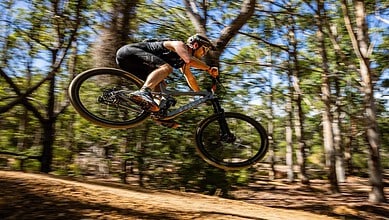 Mountain biker in mid-air jump through forest trail during a mountain biking event, showcasing skill and adventure in outdoor terrain.